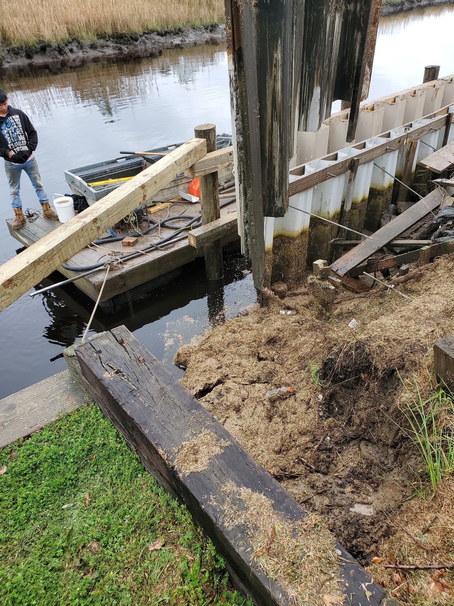 Damaged wooden dock and railing beside a canal, with a person standing by the water.