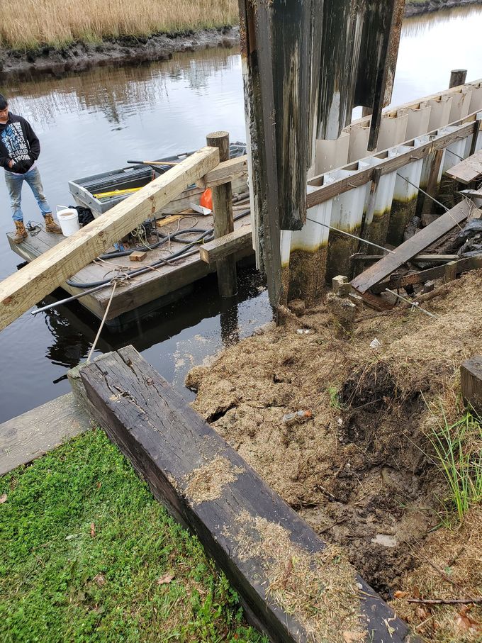 Damaged wooden dock railing beside a canal with one person standing nearby and debris in the water