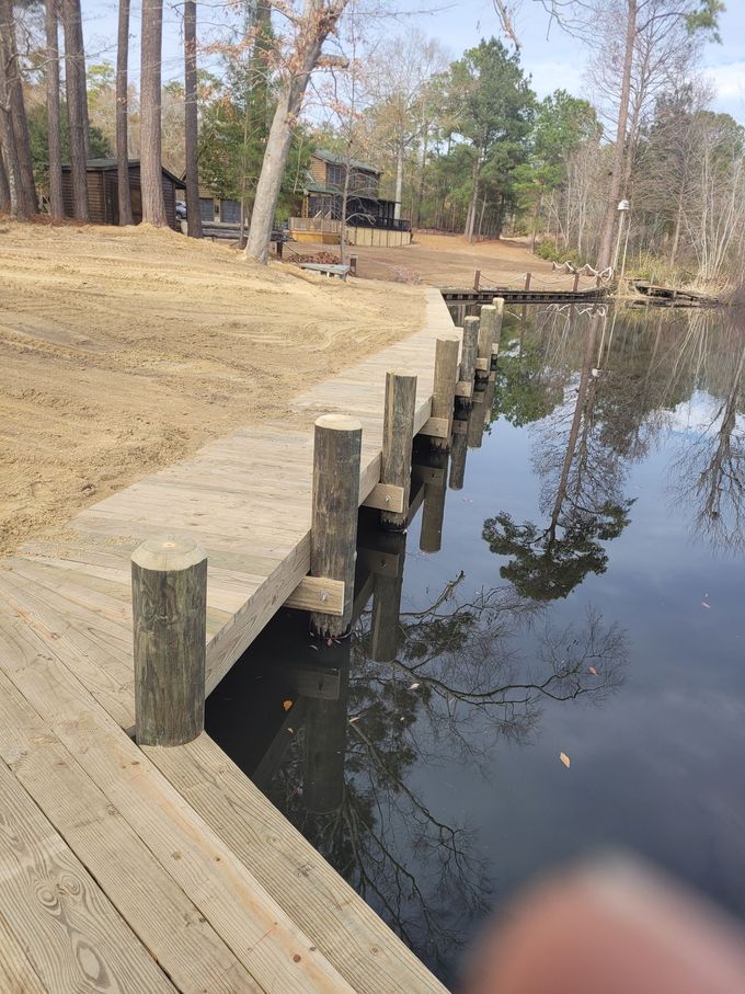 Wooden dock beside a calm creek, with trees and houses reflected in the water.