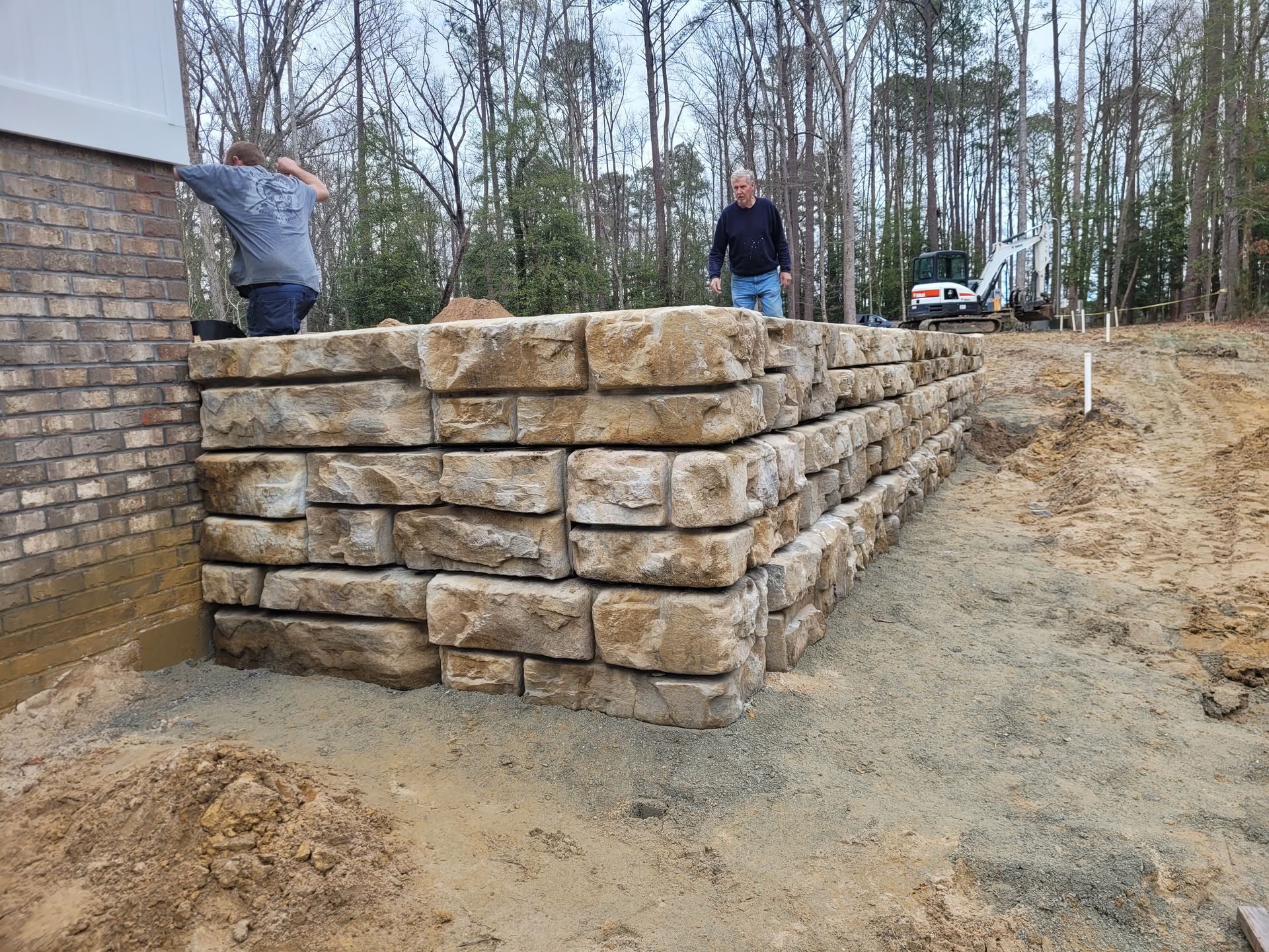Stone retaining wall under construction with two workers at a wooded building site.