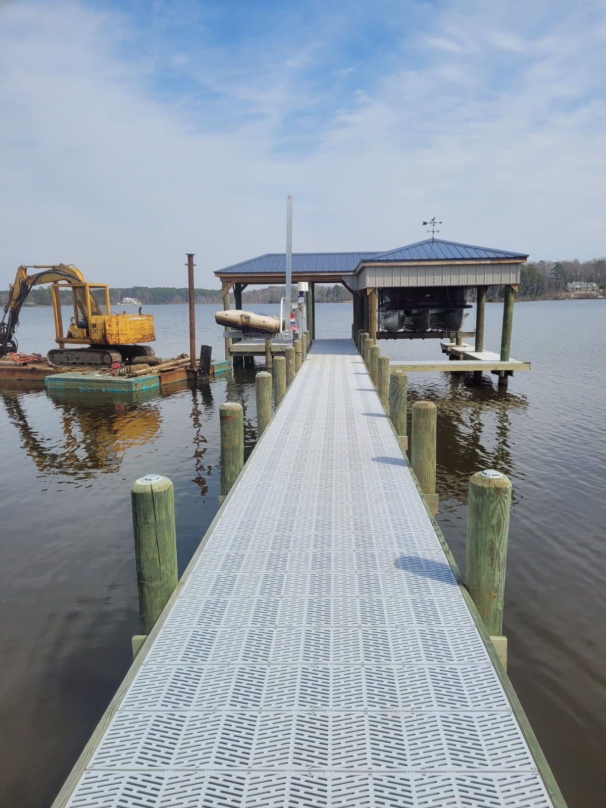 Dock leading to a blue-roofed boathouse over calm water, with a yellow excavator on the left.
