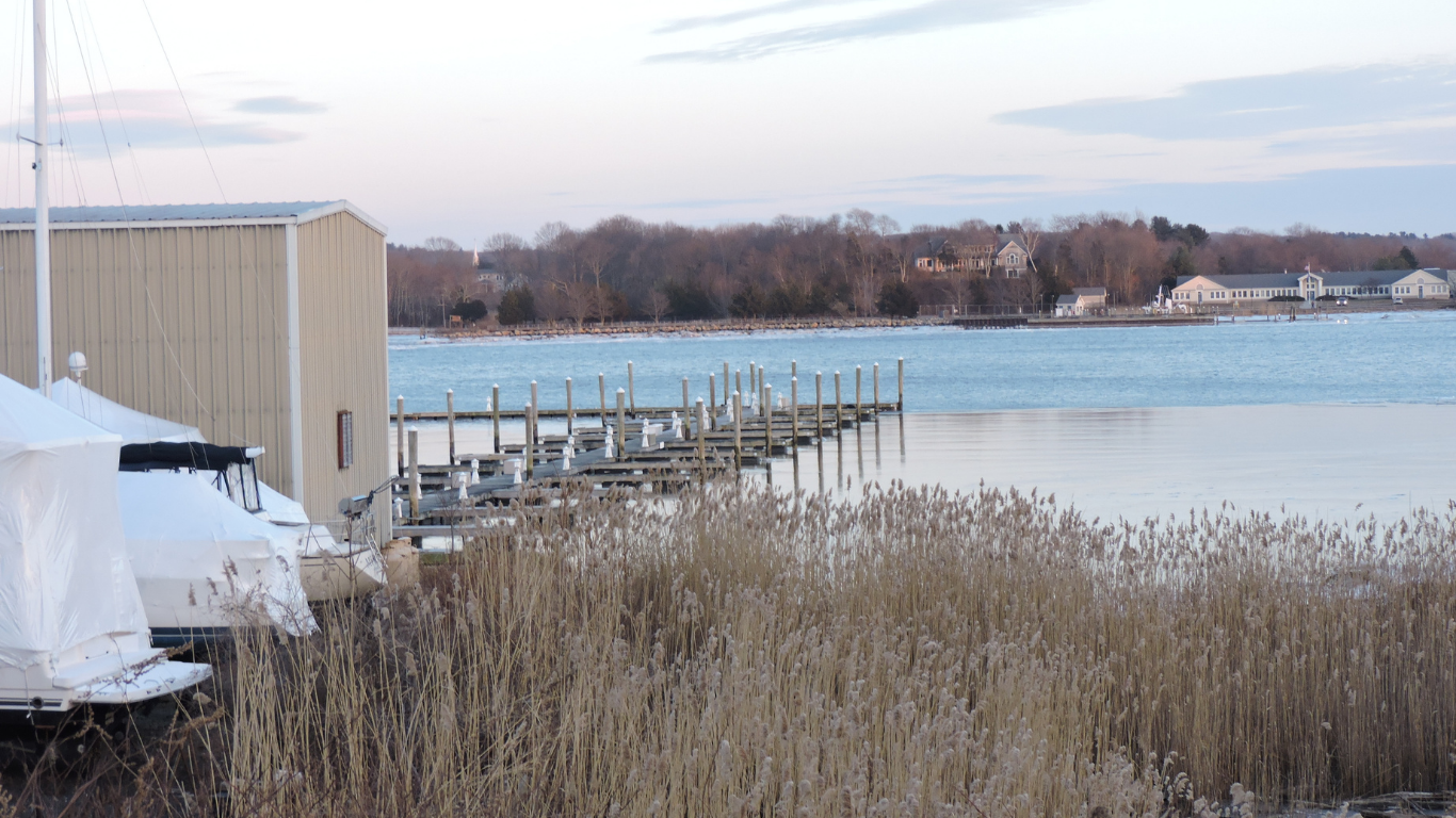 Frozen waterfront dock beside beige building, with reeds in foreground and calm water beyond