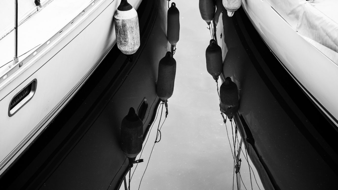Black-and-white view between two docked boats reflected in calm water.