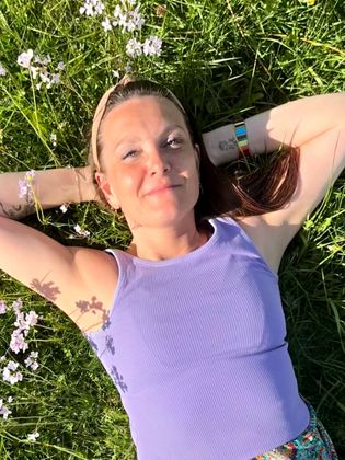 Sabine FHealS in  een paarse tanktop ligt in een zonnig, met bloemen gevuld grasveld met de handen achter het hoofd.