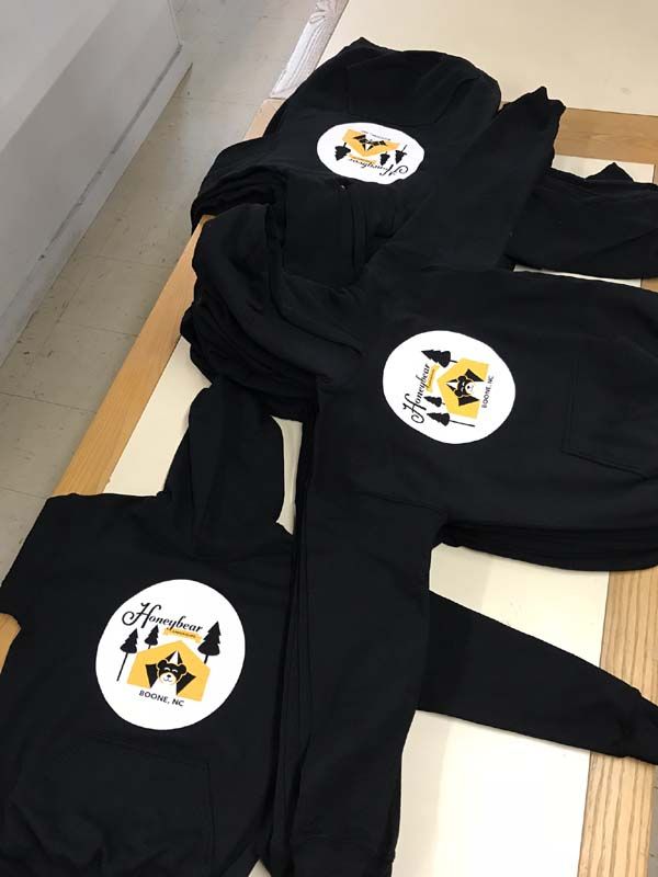 Black sweatshirts with white and yellow logo; stacked on a wooden surface.