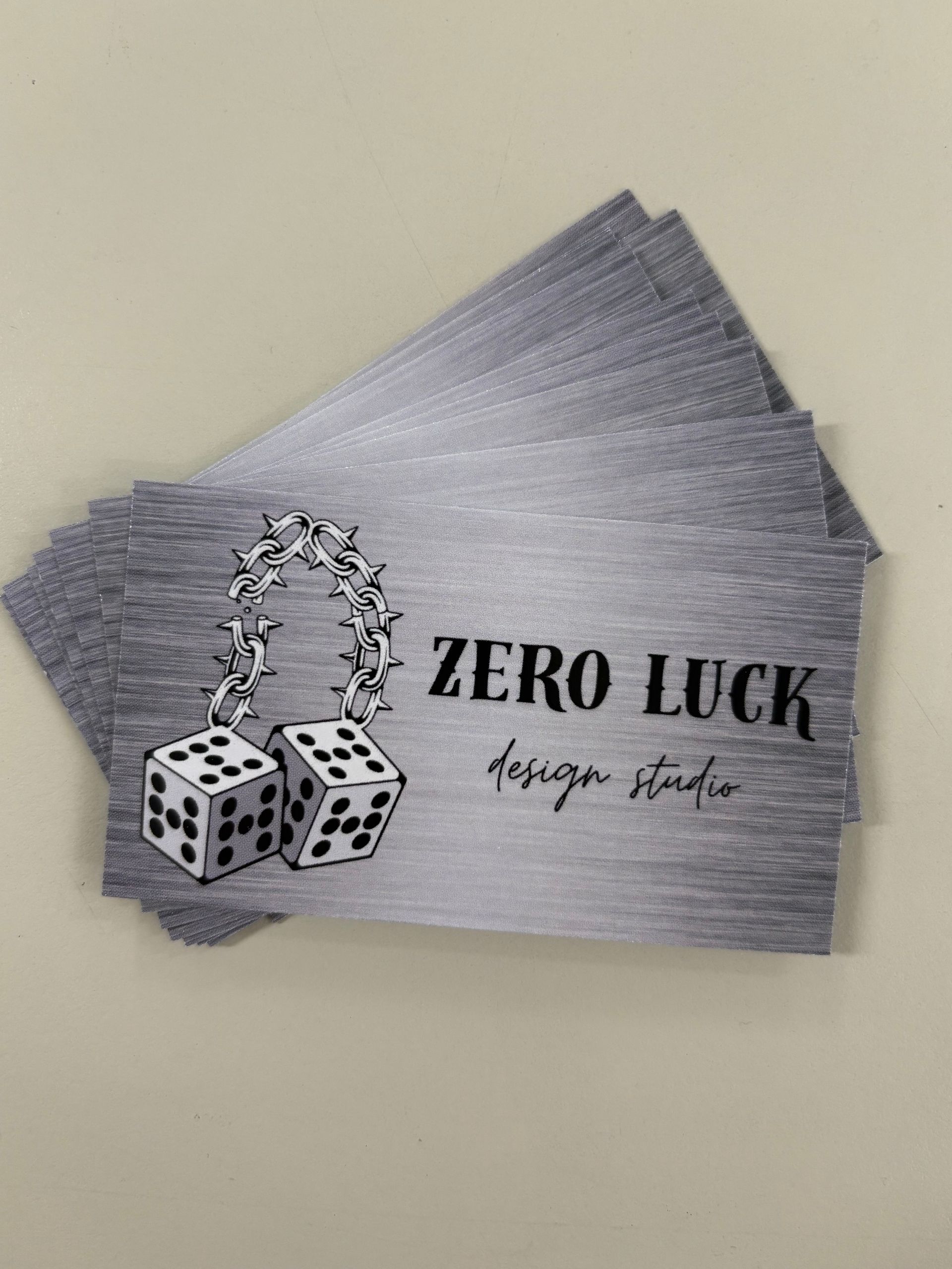 Stack of gray “ZERO LUCK” business cards with dice and a chain graphic on a beige surface