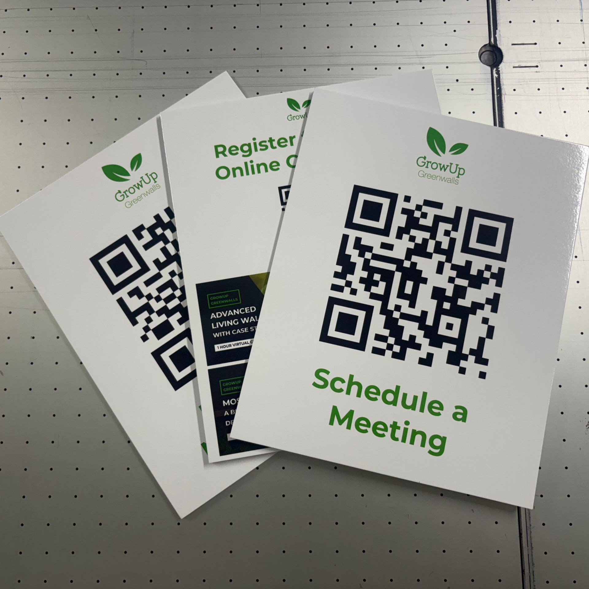 Three white QR code flyers on a pegboard, with green text including “Schedule a Meeting” and “Register online”