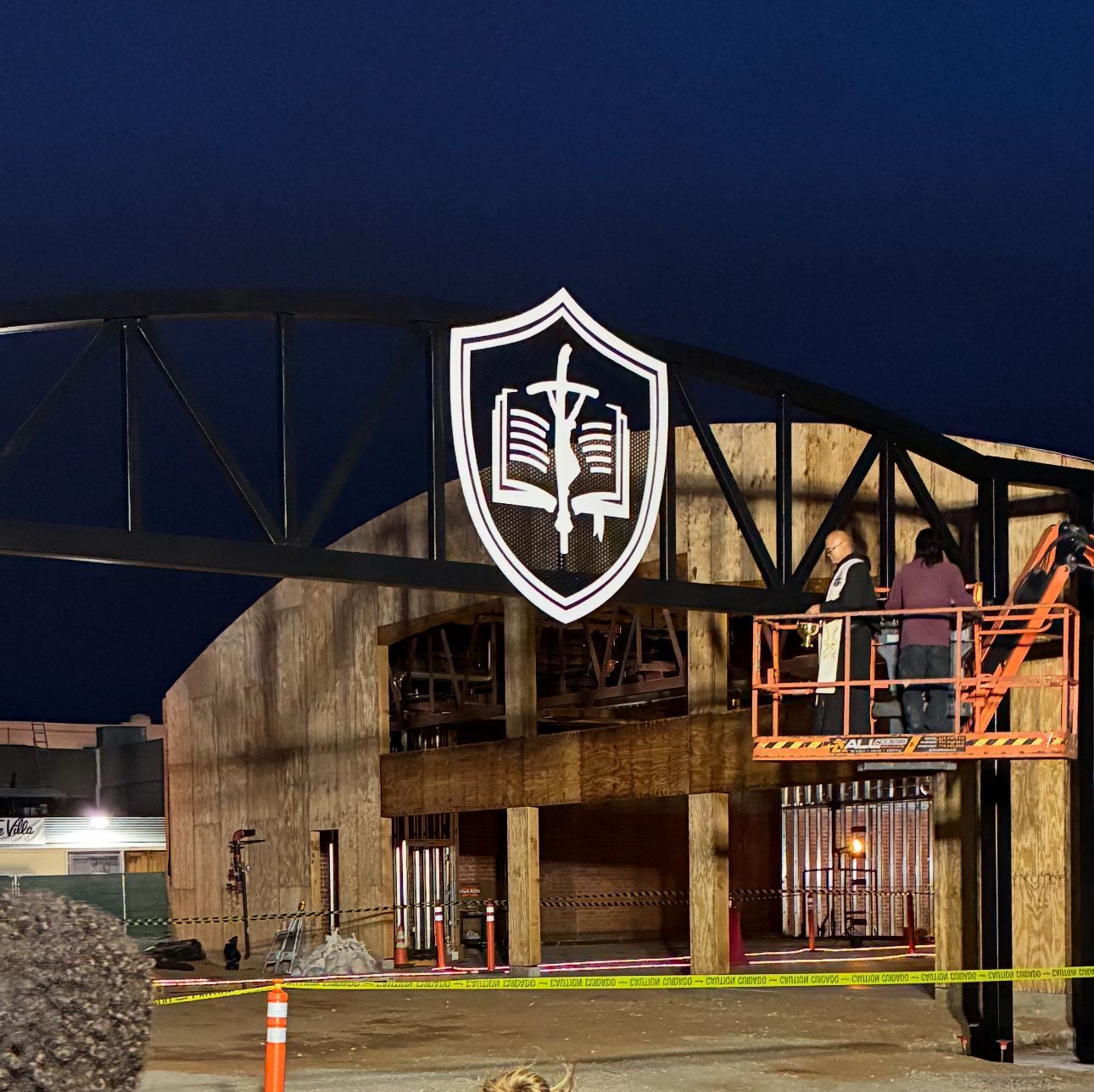 Nighttime construction site with a brick building, raised worker platform, and large shield sign hanging overhead