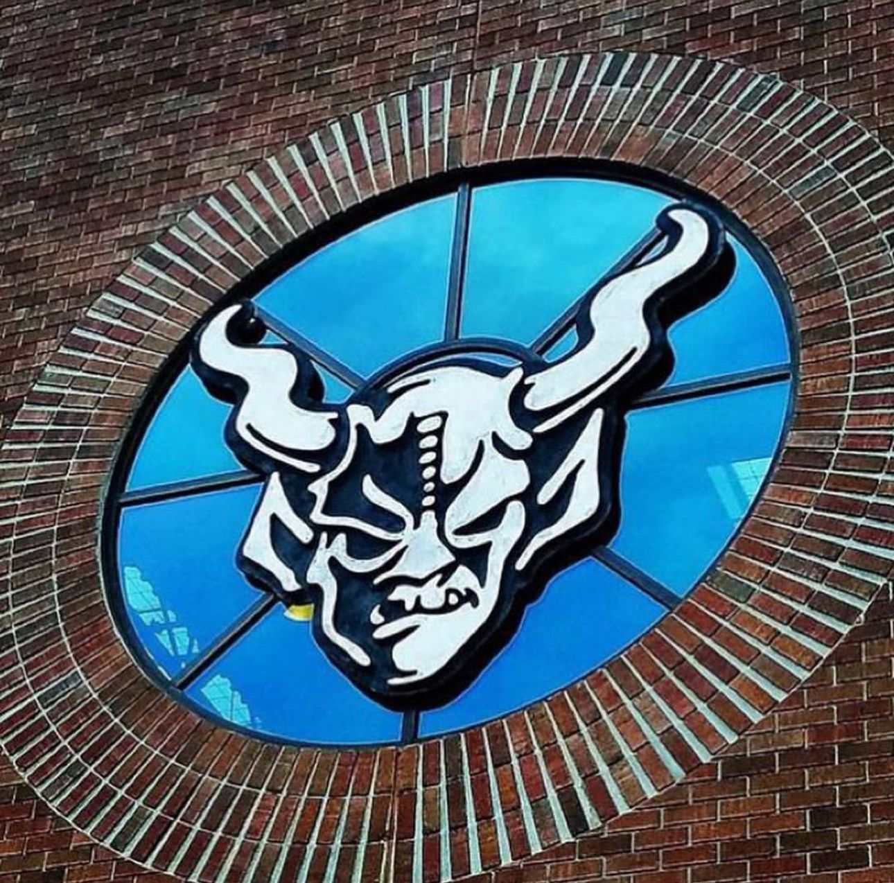 Stylized horned mascot logo in white and black on a blue circular window set in a brick wall