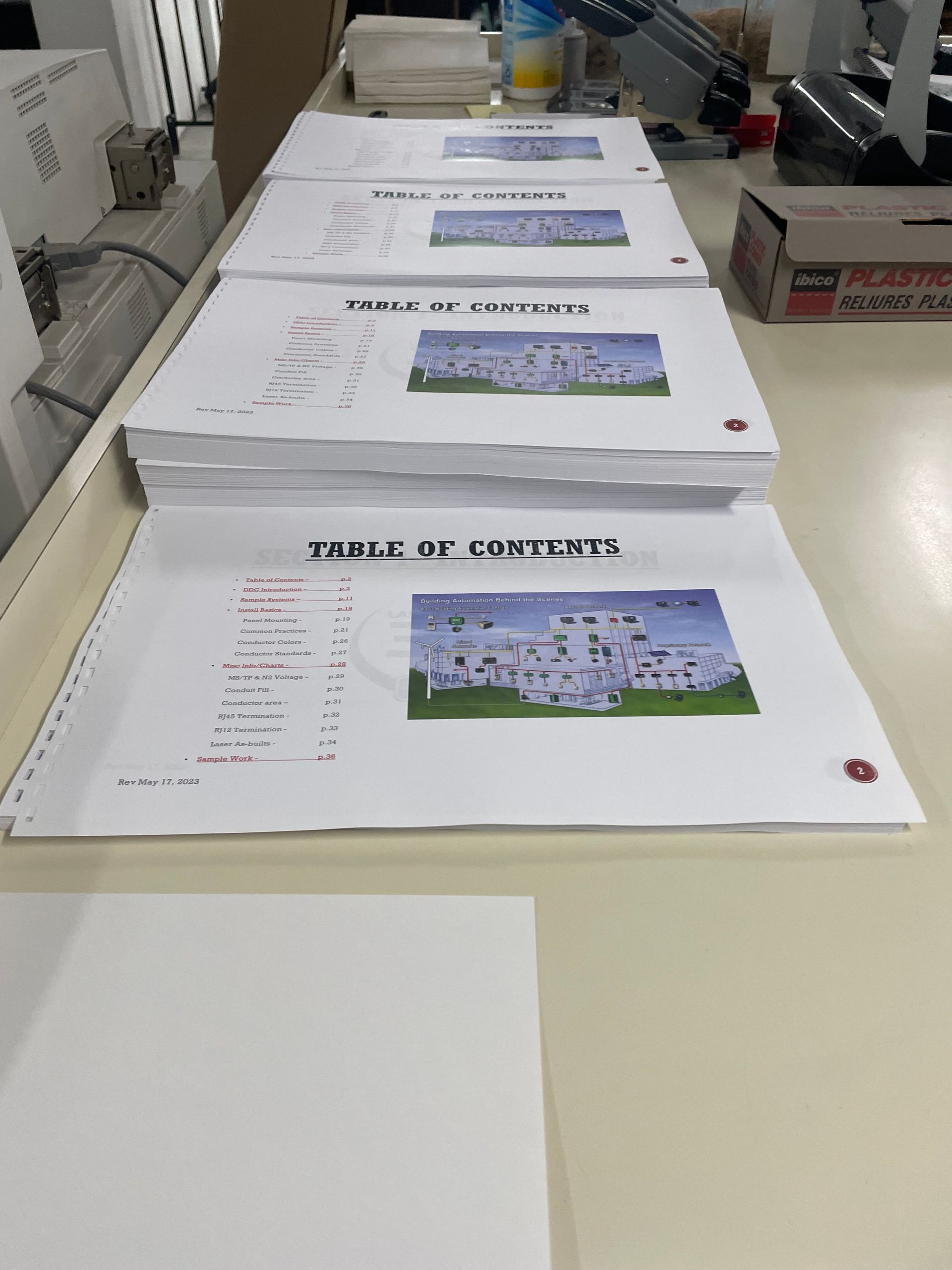 Stack of printed pages on a white table in a workshop, each showing a building photo and text.