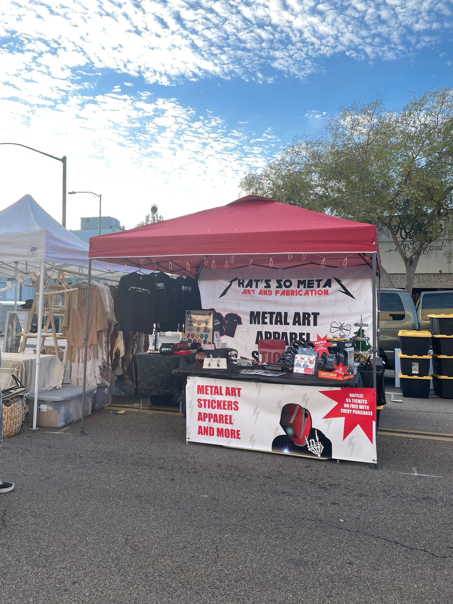 Outdoor vendor booth under red canopy selling metal art and memorial items at a market.