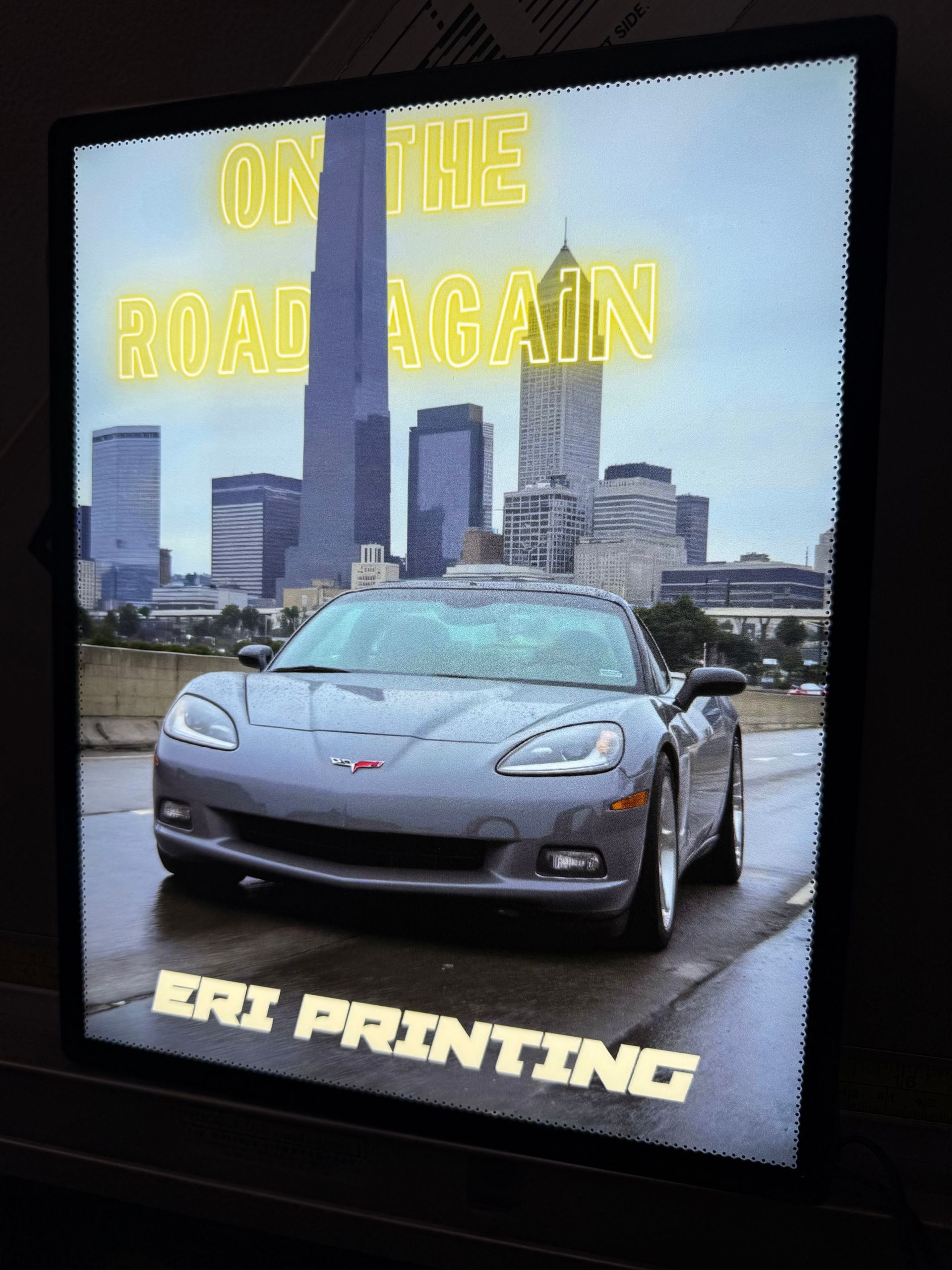 Silver sports car in front of a city skyline with yellow text “On the Road Again” and “ERI PRINTING”