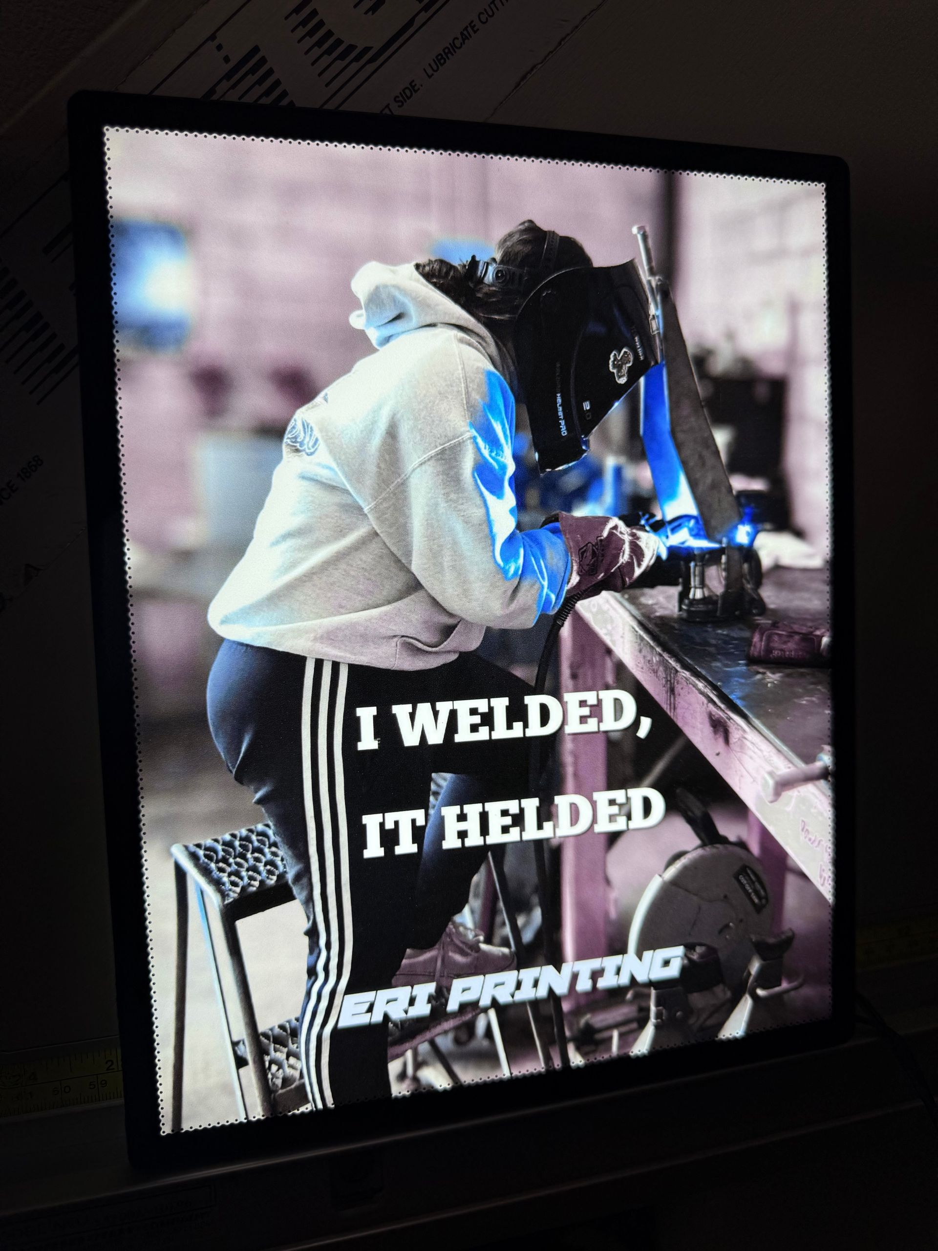 Welder working on metal with bright blue sparks, wearing a hooded jacket; poster text reads “I WELDED, IT HELDED.”