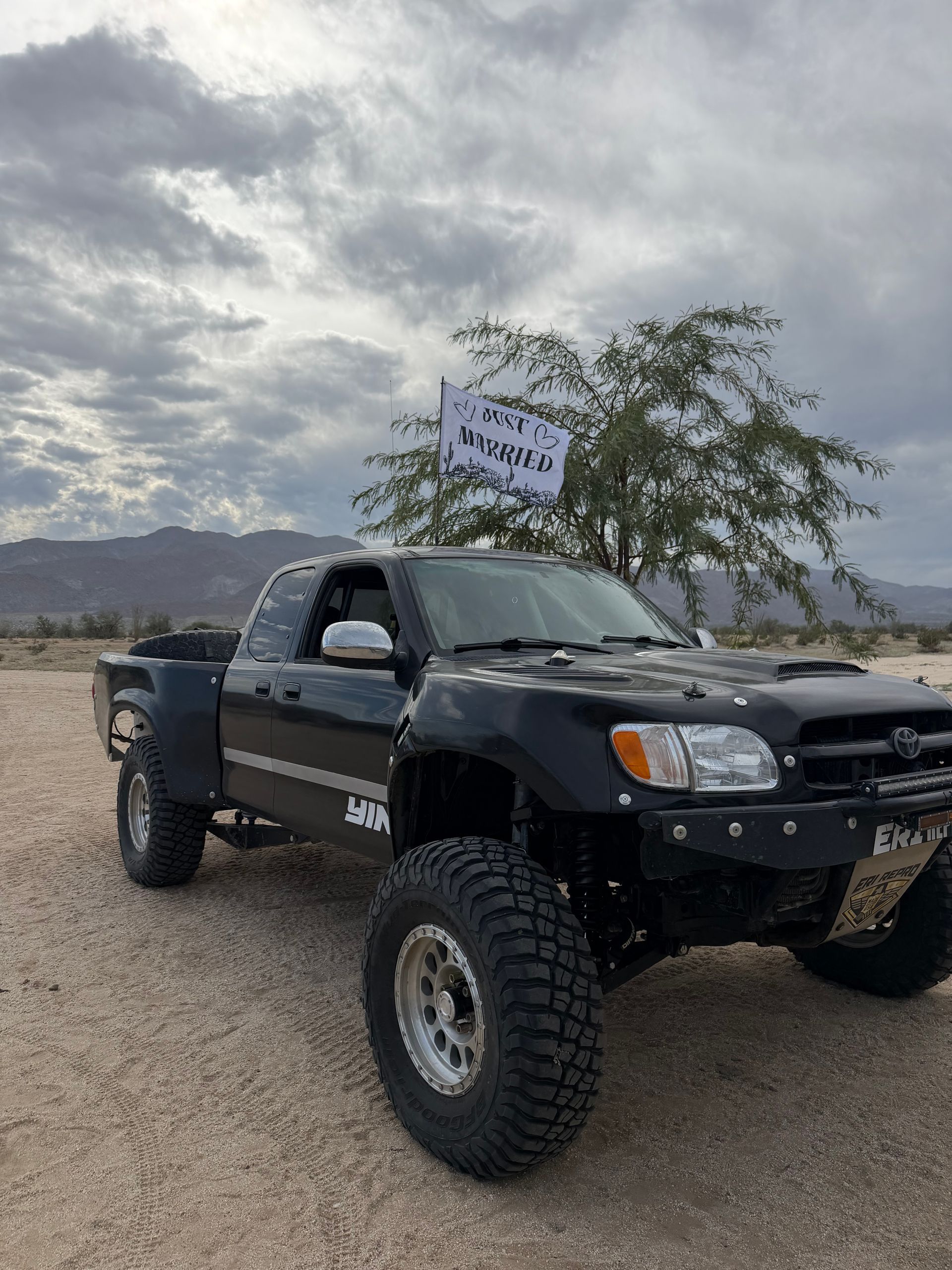 Black lifted pickup truck parked on a gravel road under cloudy skies near a sign and tree