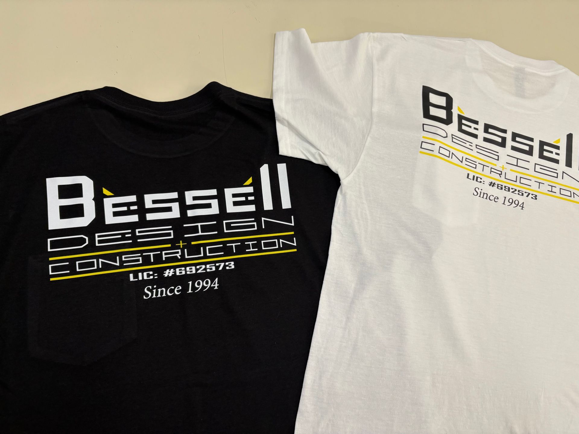 Two Bessel Construction logo T-shirts, one black and one white, laid flat side by side.