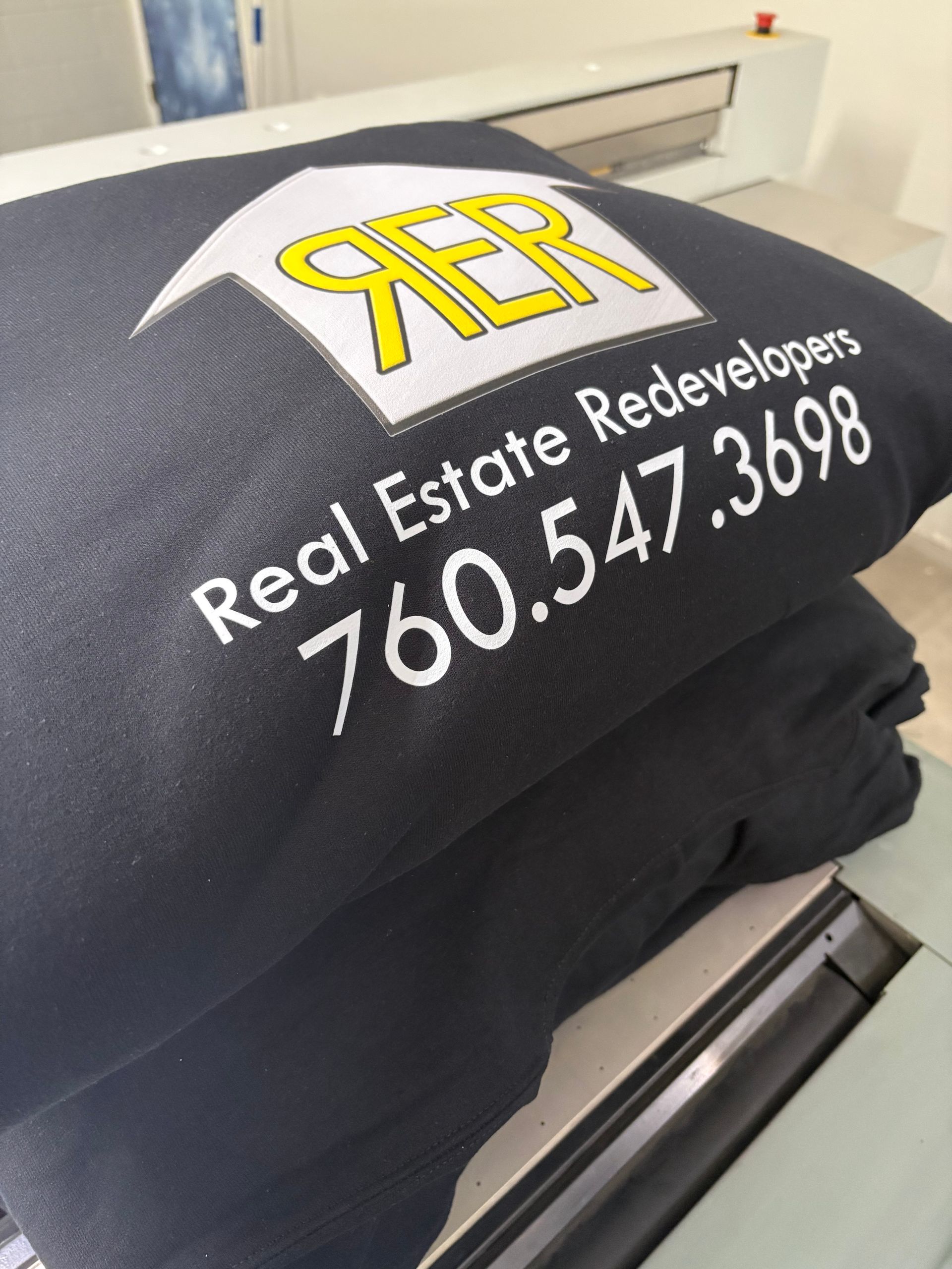 Black T-shirt with yellow “RER” logo and white text “Real Estate Redevelopers 760.547.3698”