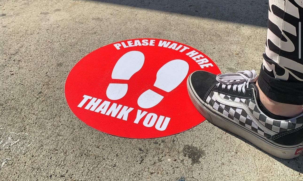 Feet standing on a red “Please wait here, thank you” floor marker on pavement.