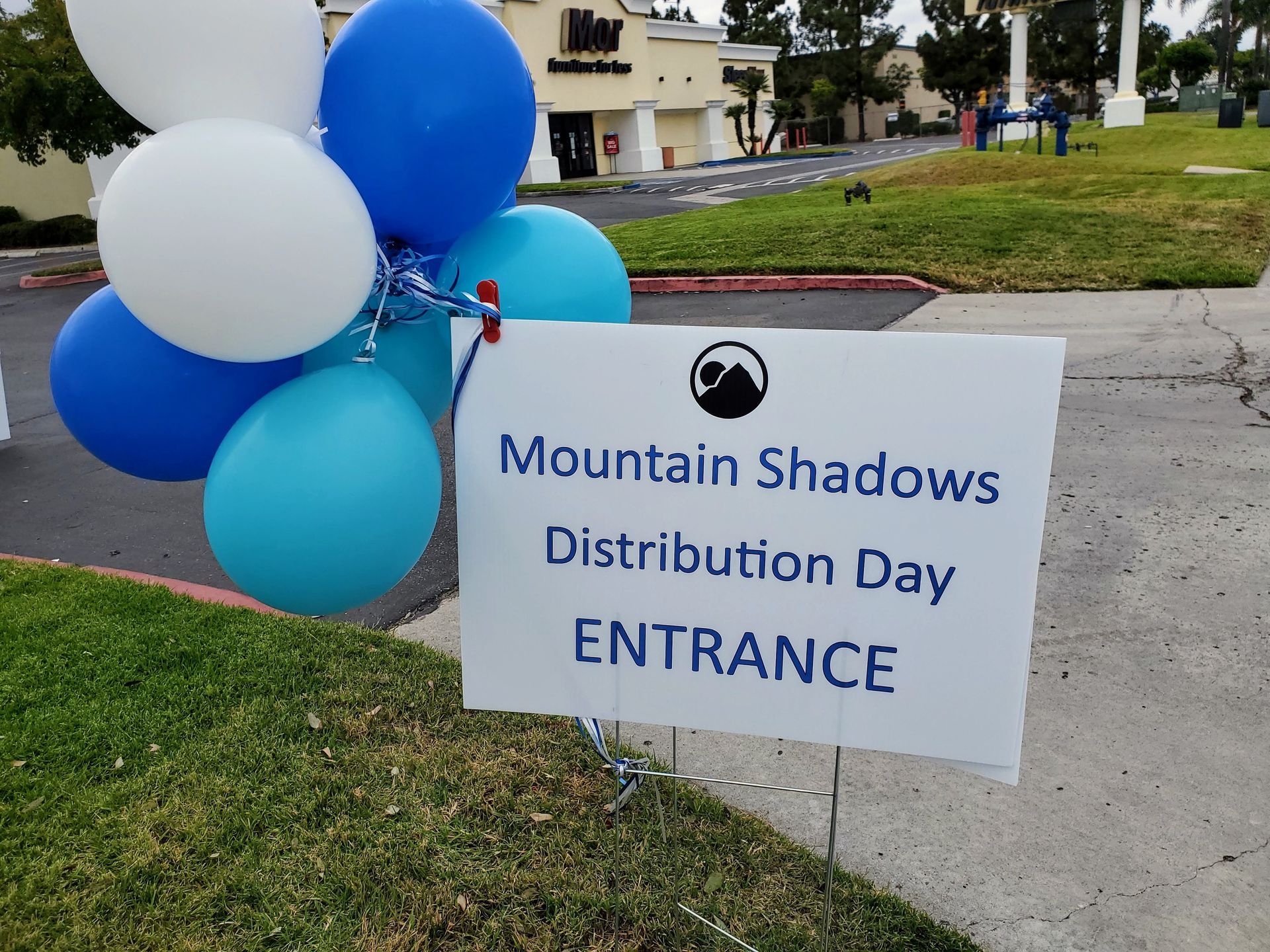 Balloon-decorated sign for Mountain Shadows Distribution Day Entrance beside a sidewalk.