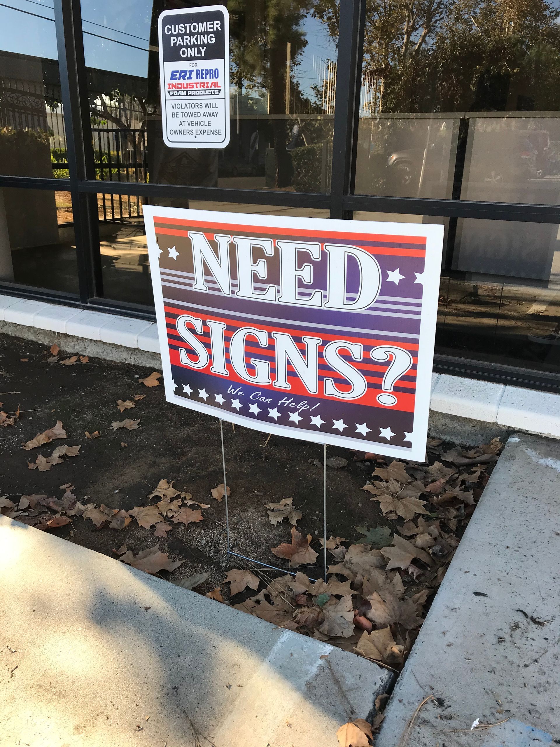 Sign in yard: “NEED SIGNS?” in red, white, and blue outside a storefront window.