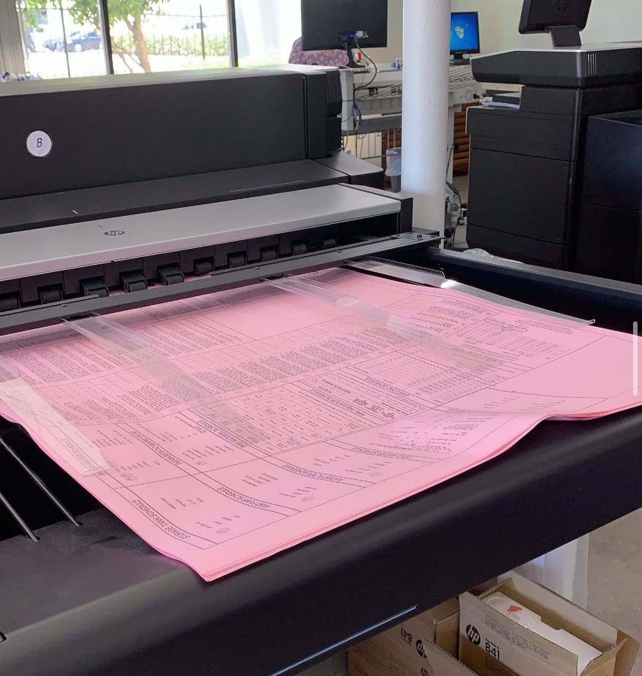 Large-format printer outputting a pink sheet with printed text in an office setting