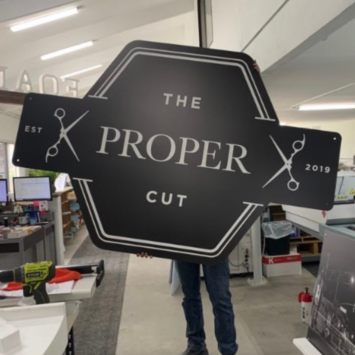 Person holding a large black barber sign reading “THE PROPER CUT” in a store.