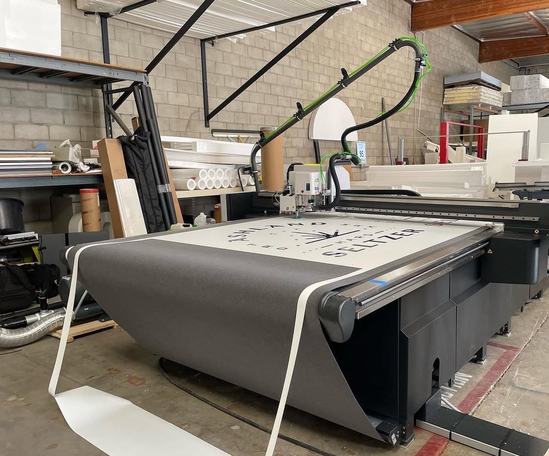Large industrial cutting machine in a workshop, with a black conveyor bed and white frame.