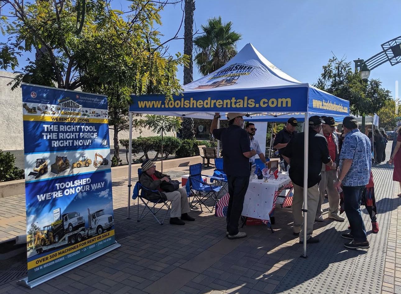 Blue canopy booth at an outdoor event with people gathered, banners, and table displays under sunny skies