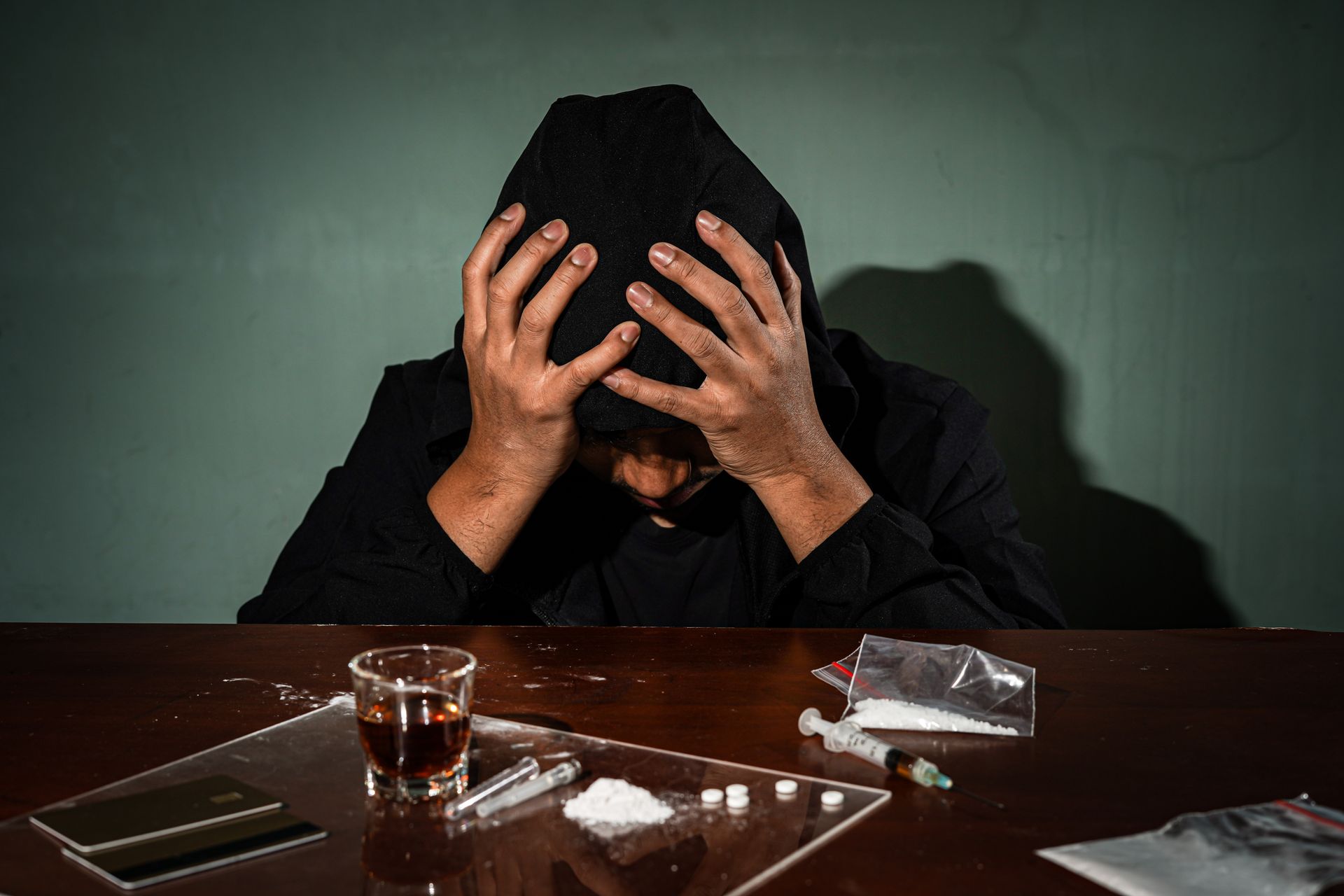 Person in a black hoodie with head in hands at a table, surrounded by a drink, cigarette, and drugs