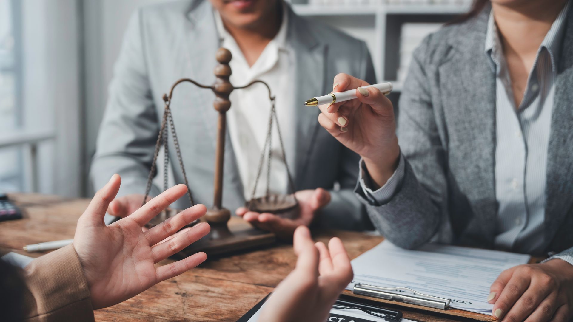 Business meeting with hands gesturing over documents, a scale of justice, and a pen in a gray office setting
