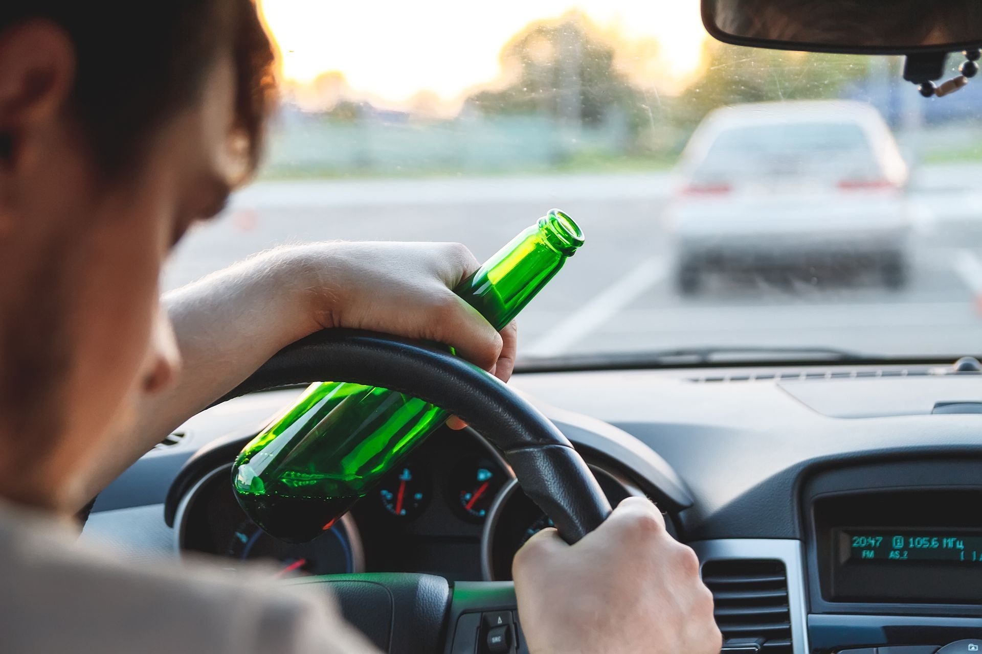Driver holding a green bottle while steering in a car, with traffic visible ahead.