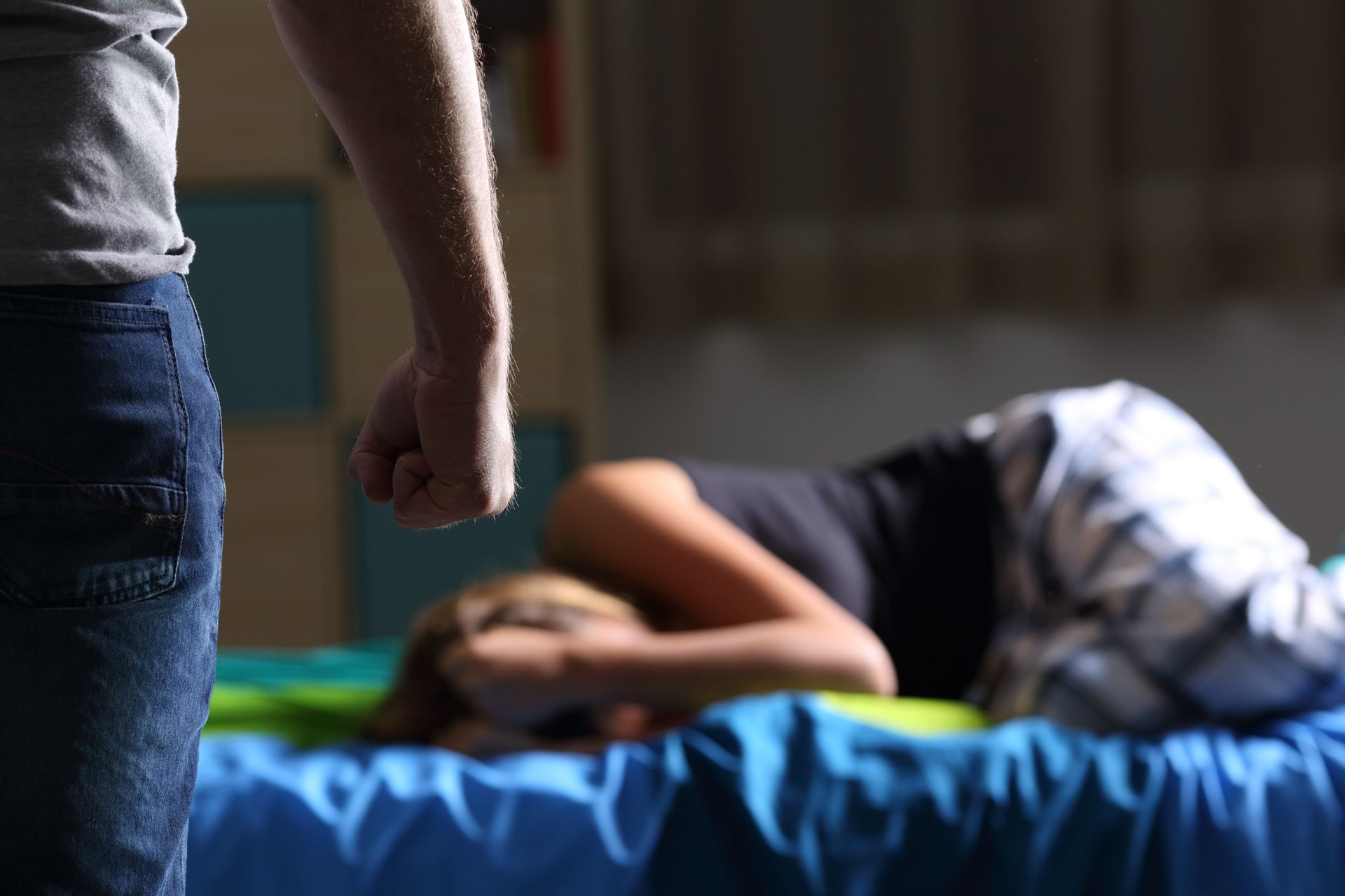 Person standing beside a bed with a person curled up under a blanket in a dim room