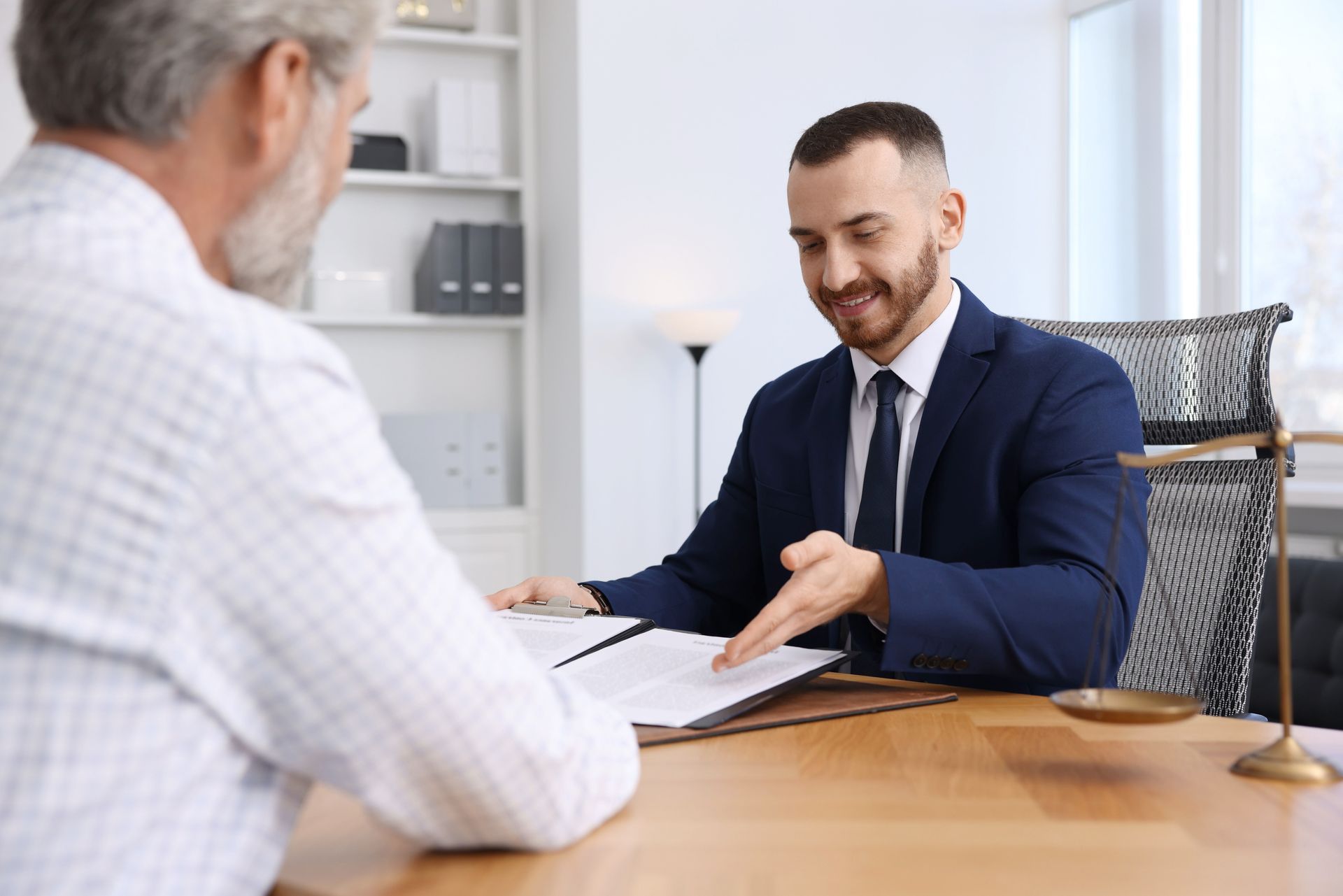 Two men discuss paperwork at a desk in a bright office, with one man pointing to a document.