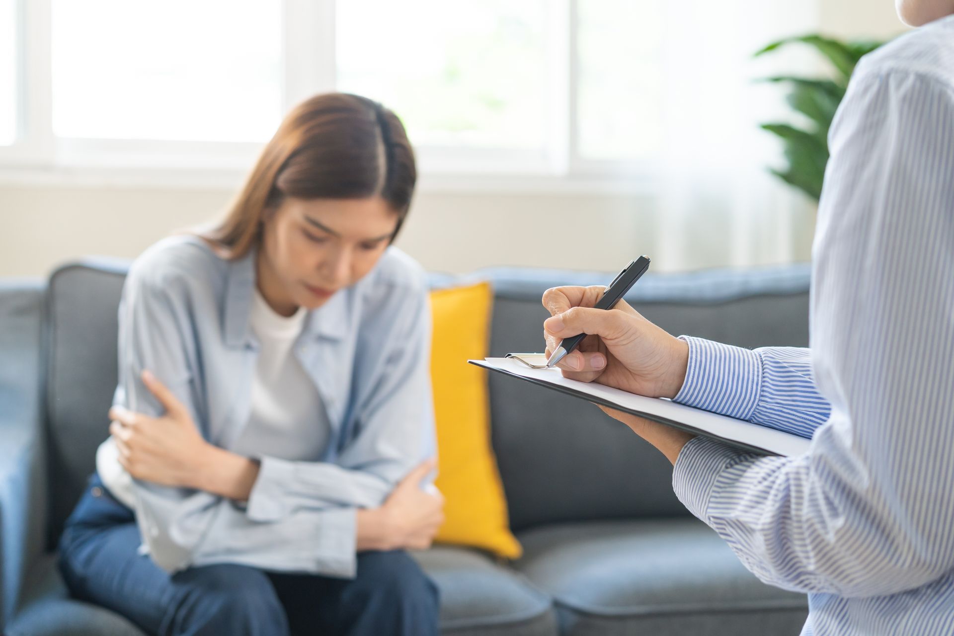 Therapist taking notes while speaking with a seated patient on a sofa in a bright living room