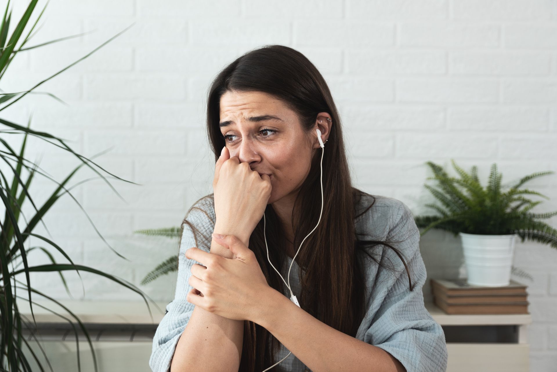 Woman sitting indoors, wiping tears and holding her arm, with plants and books in the background.