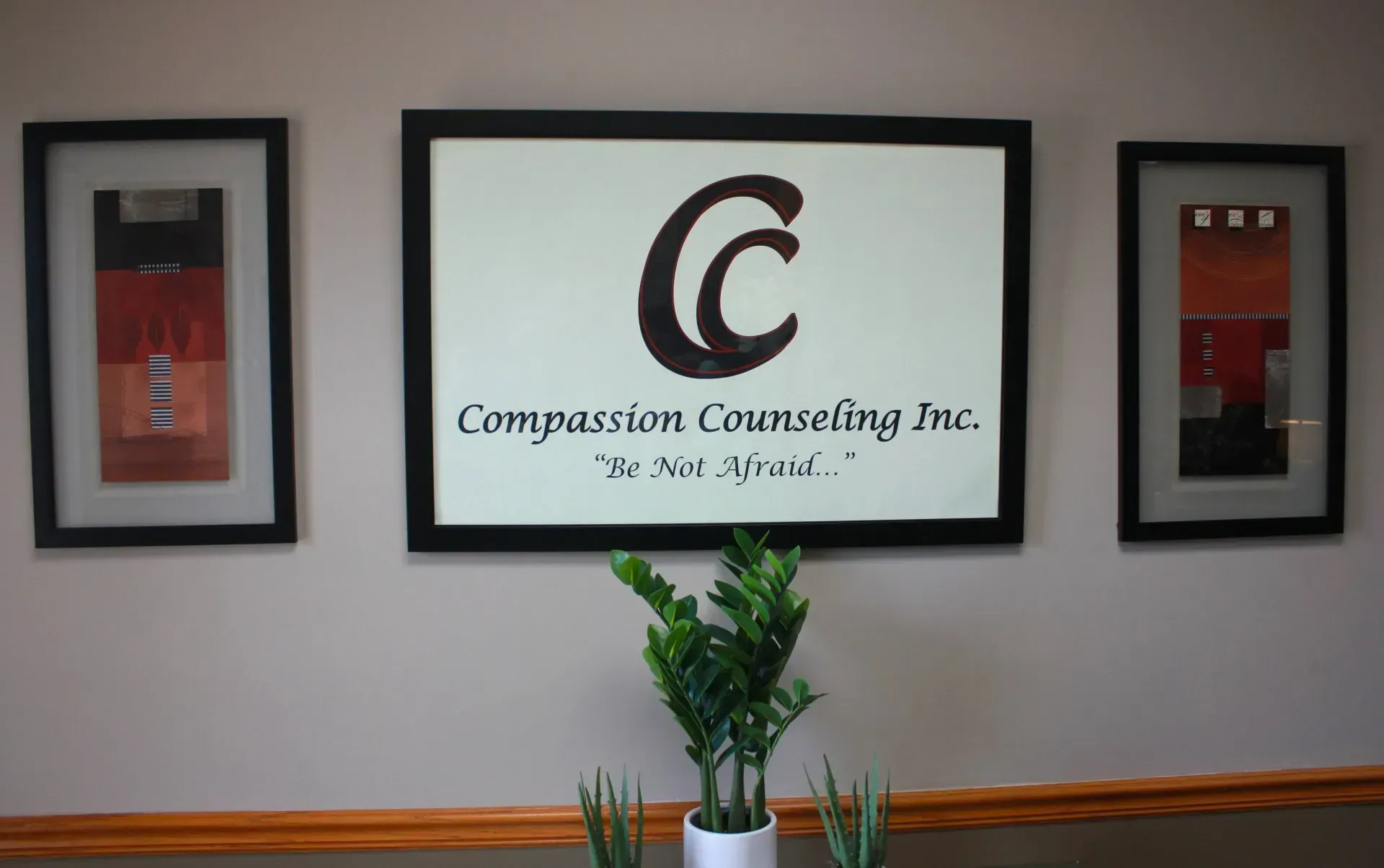 Office wall with three framed pieces, centered logo for Compassion Counseling Inc. above a plant