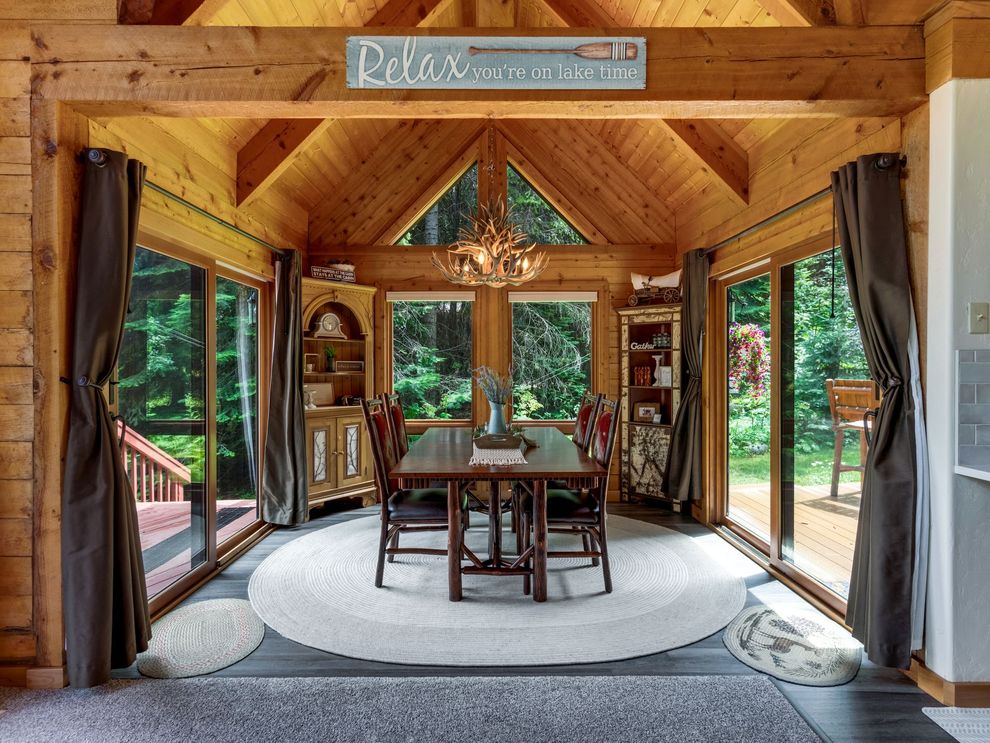 Wood-paneled dining room with large windows, round table, and chairs overlooking a forest deck