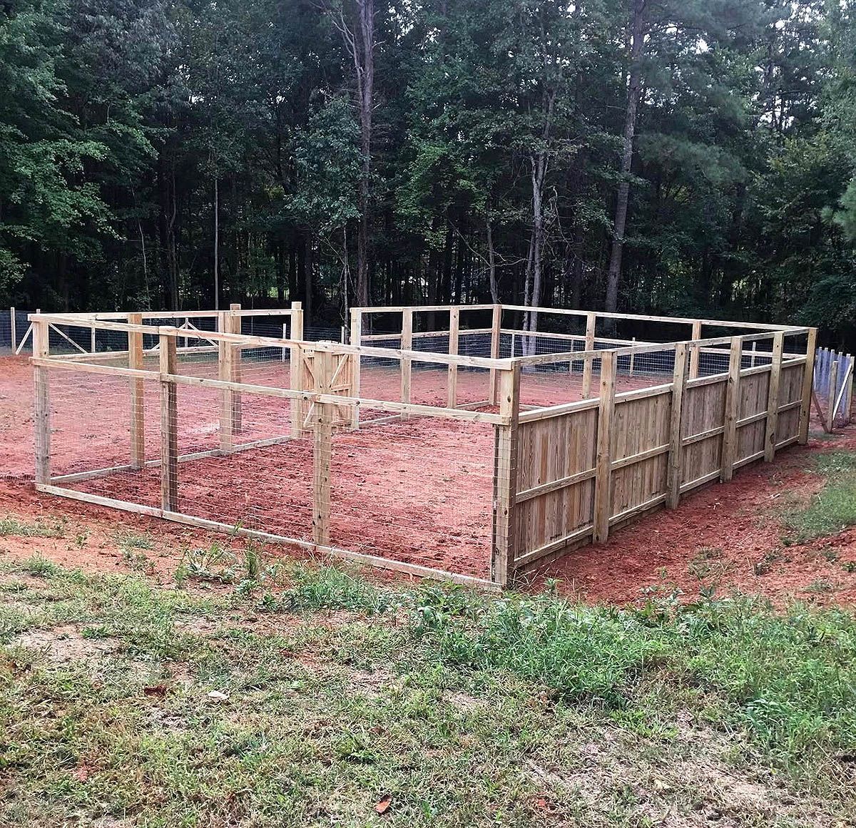 Wooden outdoor fence enclosure with posts and red dirt, set beside a wooded area.