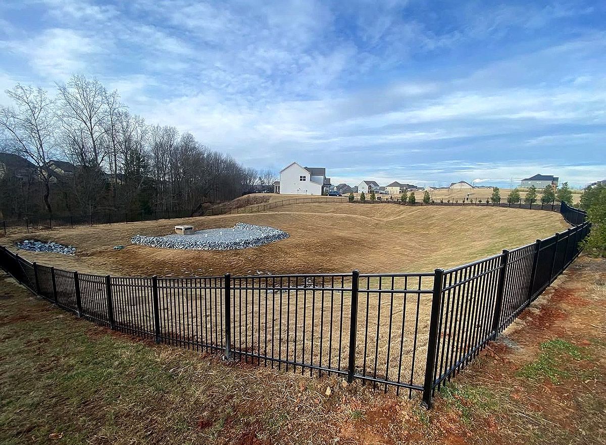 Black fenced outdoor field with a small pond, leafless trees, and houses under a cloudy sky