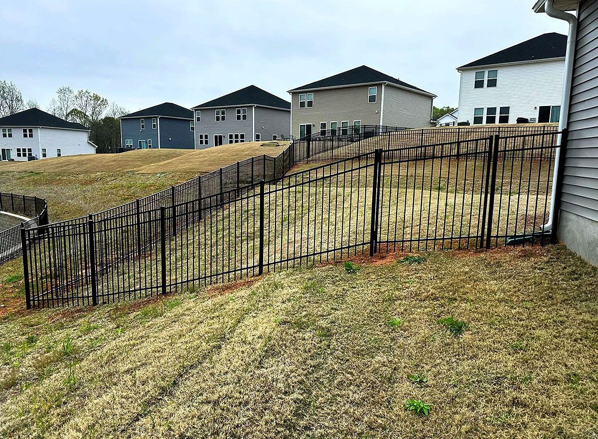 Black metal fence along a sloped grassy yard in a suburban neighborhood, with houses in the background