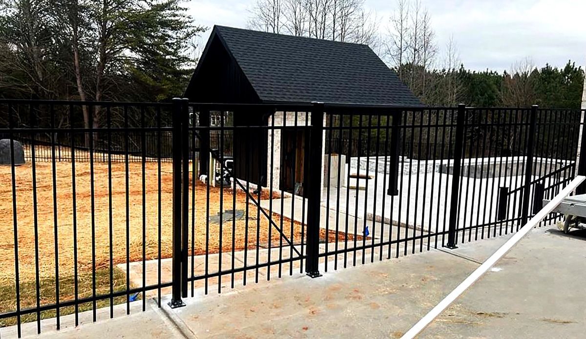 Black metal fence and gate beside a small dark shed on a concrete path, with a grassy yard and trees behind.