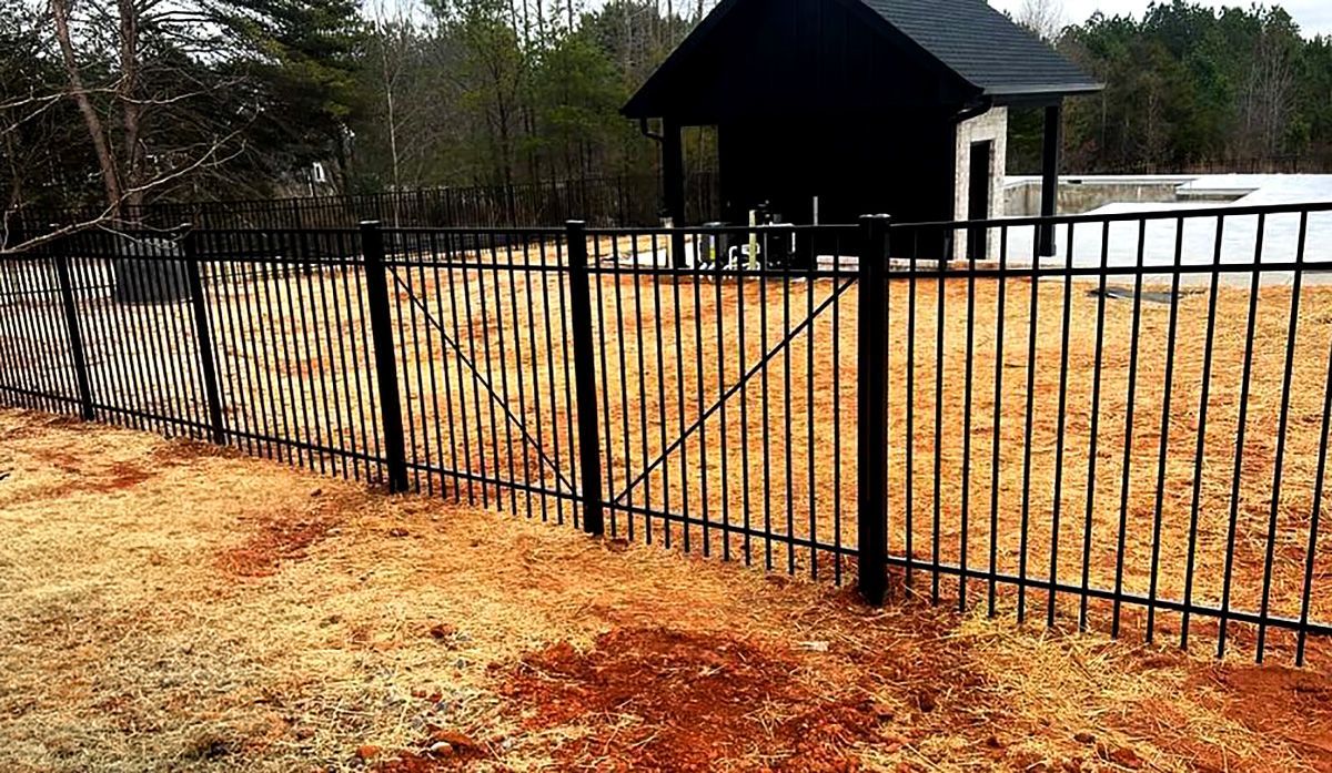 Black metal fence along a yard with a small dark shed and bare trees in the background.