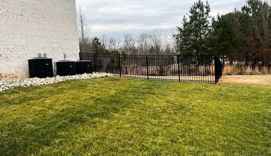 Grassy backyard beside a white house, with utility units, rocks, and a black metal fence under a cloudy sky
