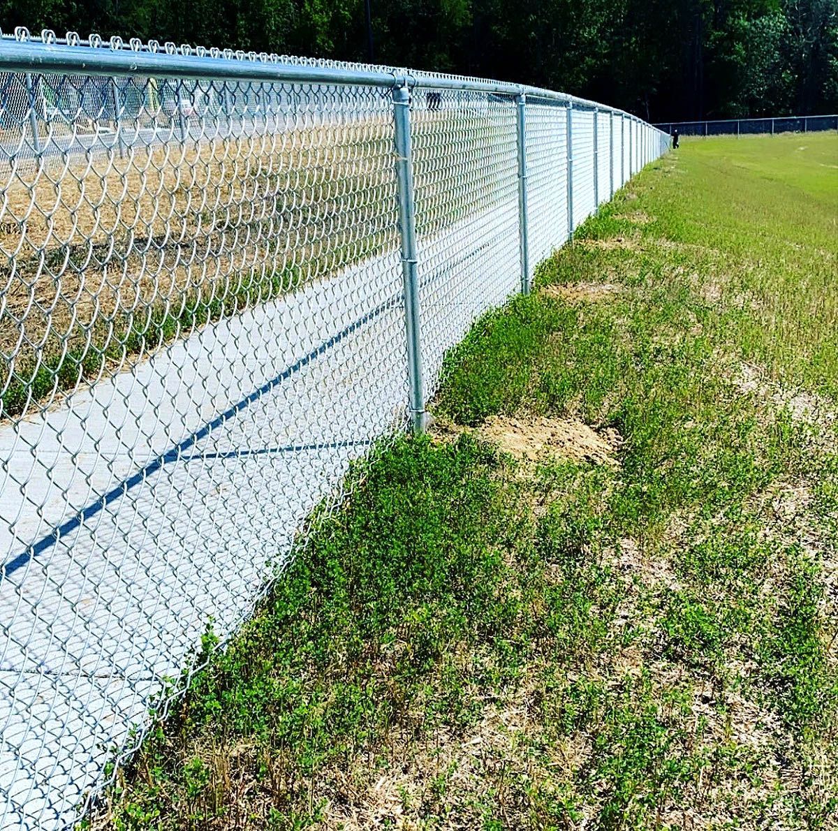 Chain-link fence along a grassy roadside embankment beside a paved path and wooded area