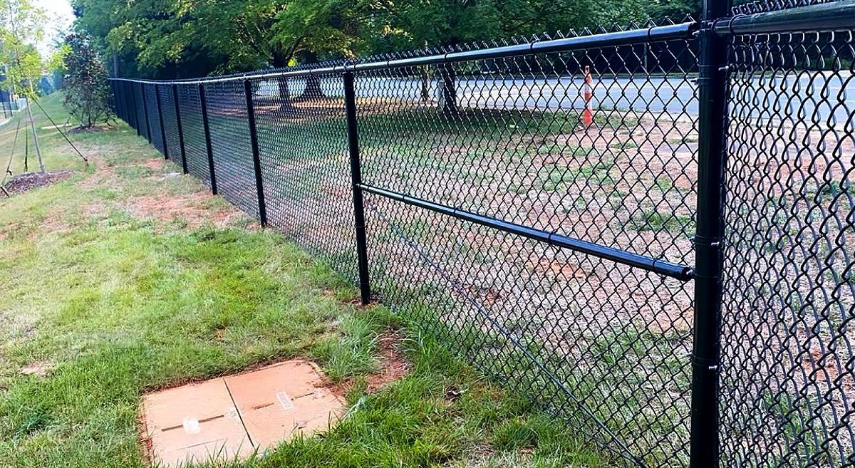 Black chain-link fence along a grassy path beside a dirt area and trees