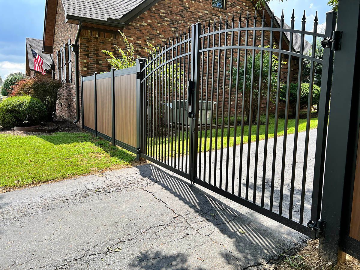 Black metal gate beside a brick house driveway, opening onto a green lawn.