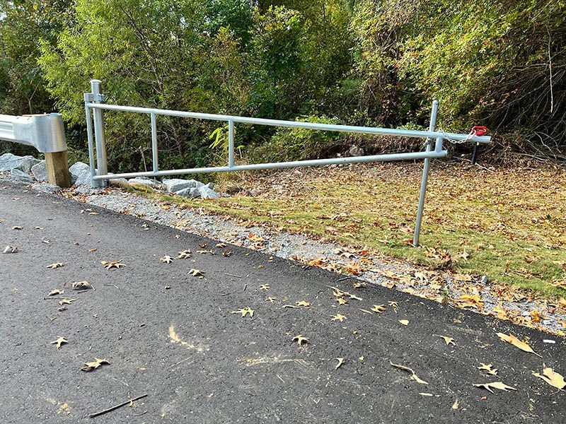 Metal barrier blocking a road beside a leaf-covered path and trees