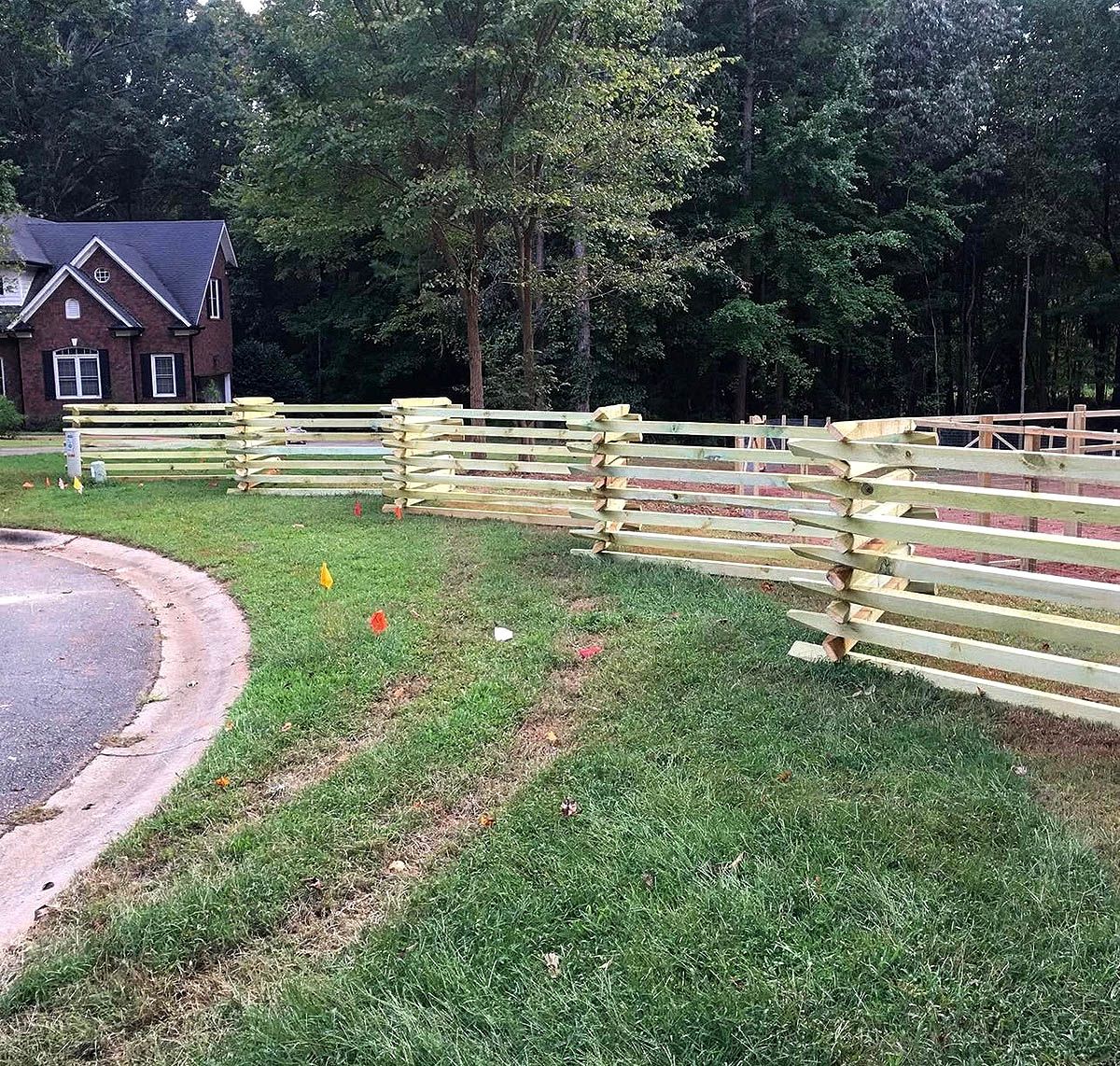 Curved wooden fence along a grassy yard beside a road, with a house and trees in the background.