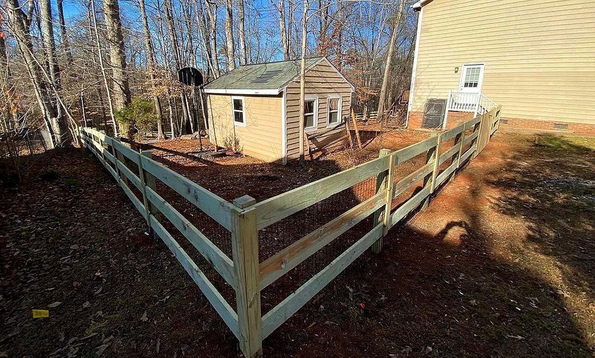 Small fenced backyard corner with a beige shed beside a house in a wooded yard