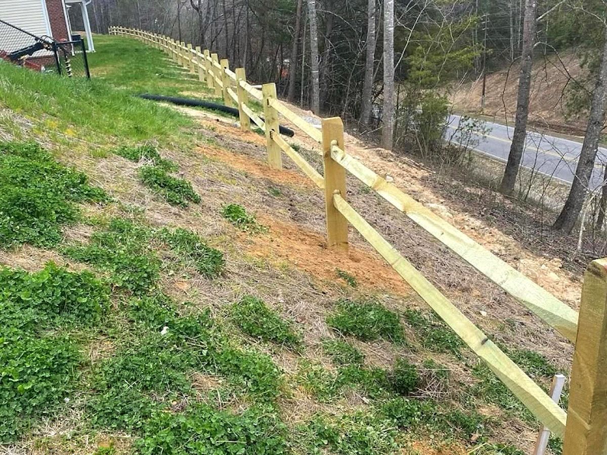 Wooden split-rail fence running along a grassy hillside beside a road and trees