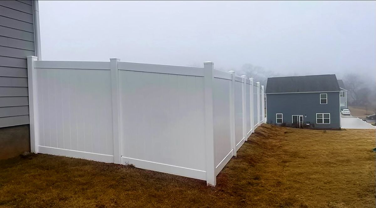 White vinyl fence beside a gray house in a foggy yard, with a second house in the distance
